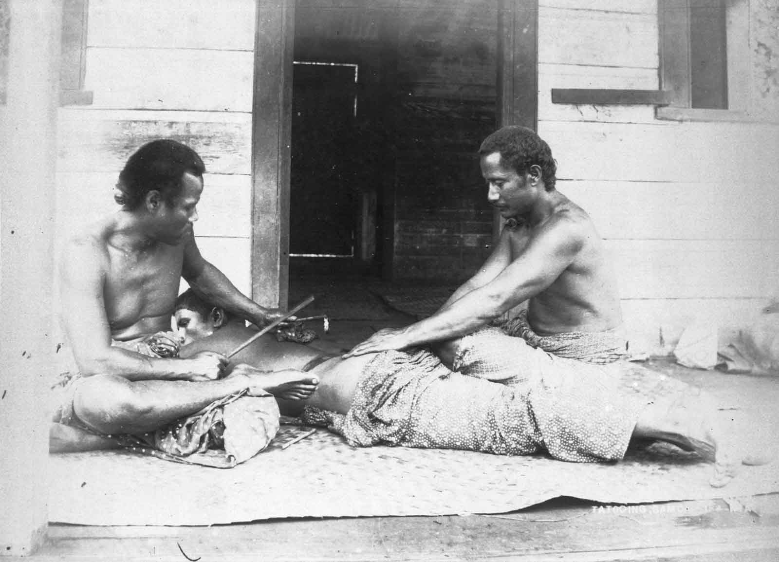 A Samoan tattooist (left), Tufuga ta tatau and assistant (right), carrying out a traditional tatau on a man's back. The tattooist uses traditional tools (1895) photographed by Thomas Andrew (1855-1939) via Wikimedia Commons